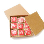 dziewięć kwadratowych pierniczków z czerwoną polewą i białymi ikonami produktów w pudełku kwadratowym, prócz tego wieczko pudełka i złota kołderka amortyzująca